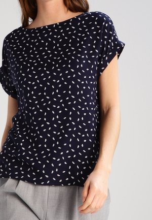 Femme portant un chemisier bleu marine à manches courtes avec un petit motif de feuilles blanches et un pantalon gris clair sur fond uni.