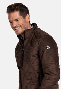 Bruine gewatteerde jas met hoge kraag, zijzakken met rits en een cirkelvormig logo-accent op de linker mouw. Zachte textuur.
