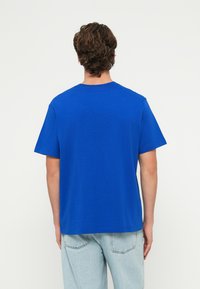 Modré bavlnené tričko s krátkym rukávom, okrúhlym výstrihom a voľným strihom, doplnené svetlými džínsami. Bez viditeľných vzorov alebo ozdôb.