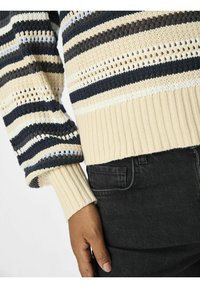 Strikket sweater med creme, marineblå og sorte vandrette striber. Ribbet ærmer med åbent vævdet detaljer, parret med mørke jeans.