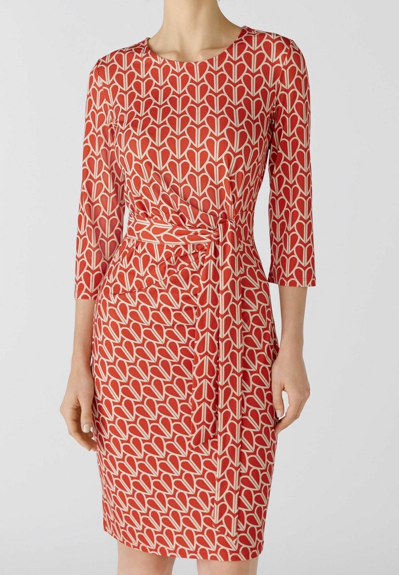 Robe rouge avec un motif de feuilles en crème, coupe ajustée, manches trois-quarts et taille ceinturée pour définir la silhouette.