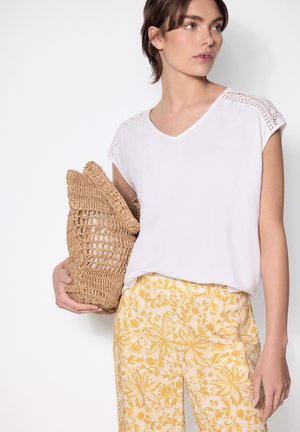 Donna con top bianco a maniche corte e pantaloni gialli floreali che tiene una grande borsa intrecciata marrone chiaro su uno sfondo bianco semplice.