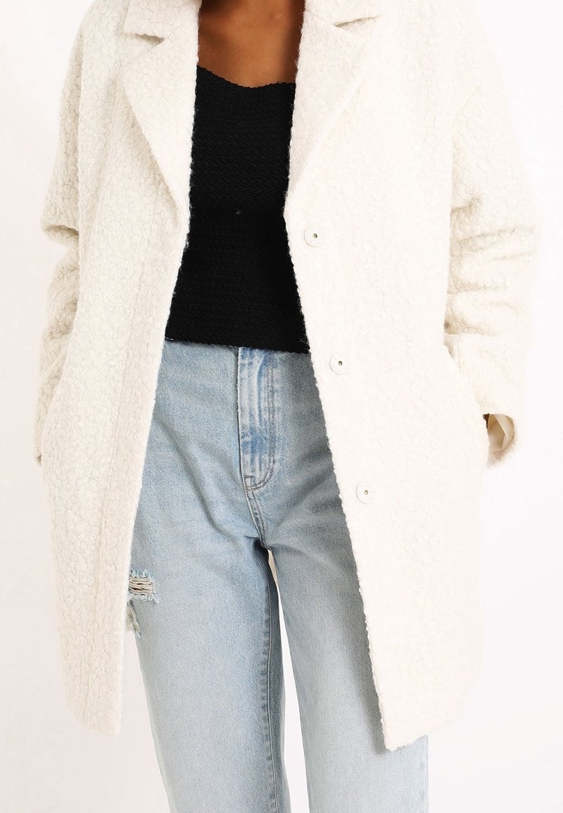 En krämfärgad, texturerad oversized rock med stora slag, framknappar och sidofickor, bärs över en svart topp och ljusa denimjeans.