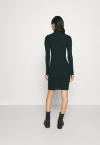 Tummaanvihreä ribbineulottu mekko korkealla kauluksella ja pitkillä hihoilla, vartalonmyötäinen siluetti, päättyy reisien puoliväliin, yhdistetty mustiin nilkkureihin.