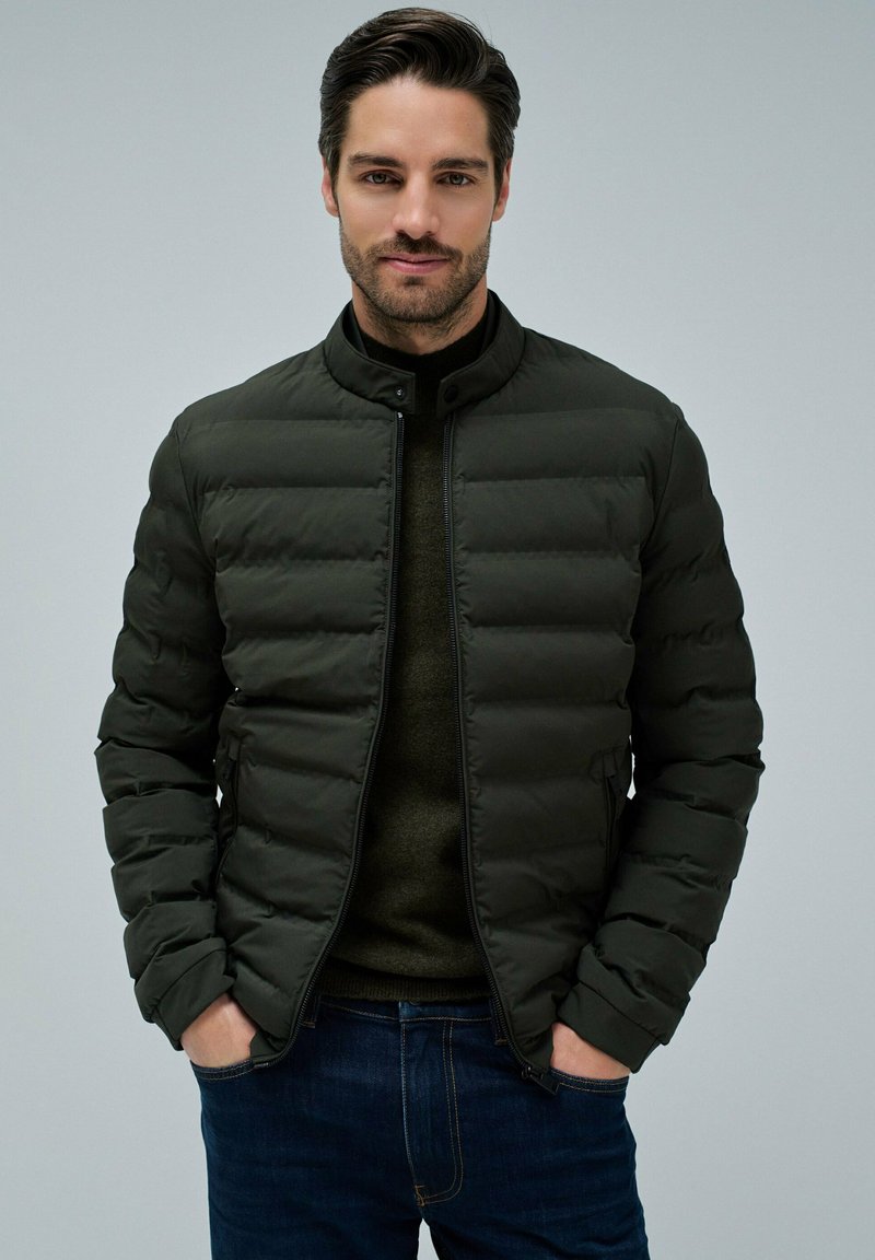Hombre con cabello oscuro que lleva una chaqueta acolchada negra sobre un suéter verde oscuro y jeans azules, con las manos en los bolsillos, fondo neutral.