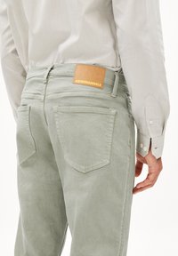 Pantalon en velours côtelé vert clair avec deux poches arrière et une étiquette en cuir portant la mention "ARMEDANGELS". Le tissu a une texture douce et des côtes subtiles.