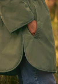 Žalias švarkas su lygiu, lengvu audiniu, turintis kišenę ir apvalintą kraštą, papildytas toniniais siūlais.