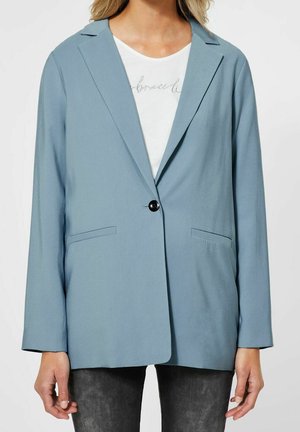 Frau trägt einen hellblauen Blazer mit einem Knopf über einem weißen T-Shirt mit Grafik und schwarzen Hosen, steht vor einem schlichten Hintergrund.