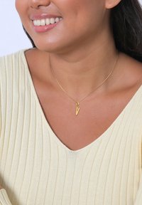 Goldene Halskette mit einem einzigartigen, V-förmigen Anhänger, getragen über einem gerippten, hellgelben Oberteil. Der Hintergrund ist neutral und übersichtlich.