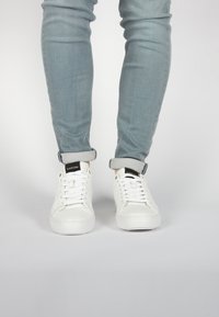 Sneakers bianchi con una texture in pelle liscia, punta rotonda e dettagli neri a contrasto sul tallone e sulla linguetta, abbinate a jeans azzurri chiari.