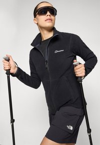 Giacca in pile nera con zip e dettagli blu, abbinata a pantaloni corti scuri. Bastone da trekking nero tenuto nella mano destra, occhiali da sole oversize indossati.