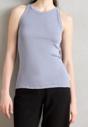 Vrouw in een lavendelkleurig mouwloos shirt en zwarte broek tegen een grijze achtergrond.