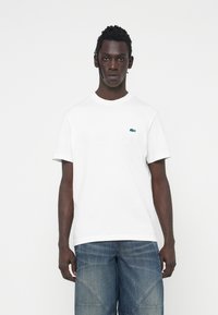 Camiseta blanca de algodón con cuello redondo y mangas cortas, que presenta un pequeño logo verde de Lacoste en el lado izquierdo del pecho. Combinada con pantalones cortos de mezclilla.