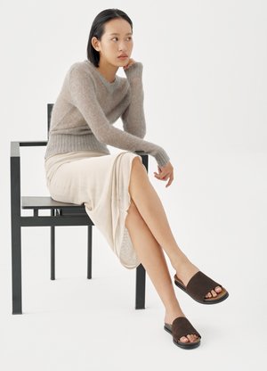 Brune ruskindsklip-sandaler med et minimalistisk design, kombineret med en lys beige nederdel til knæet og en blød grå sweater, siddende på en sort stol.