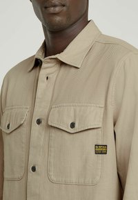 Beige skjorta med knappar, två bröstfickor, mörka knappar och en G-Star Originals-etikett på vänster ficka. Texturerat tyg med en klassisk design.