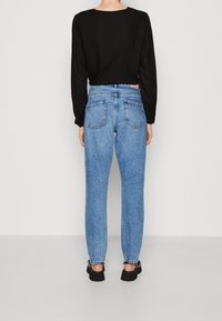 En person står vänd mot en vägg, iklädd en svart långärmad topp, högmidjade ljusblå jeans och svarta skor med tjocka sulor.