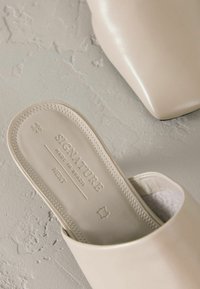 Beige nahkakengät, joissa on sileä pinta, muotoillut pohjalliset ja kohokuvioidut yksityiskohdat, mukaan lukien "SIGNATURE MADE IN BRAZIL."