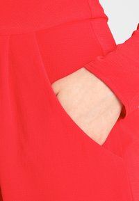 Le vêtement rouge présente une texture lisse et une poche latérale, avec un gros plan sur une main reposant à l'intérieur, montrant la flexibilité du tissu.