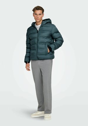 Giacca a vento teal con cuciture orizzontali, chiusura con zip e cappuccio. Il modello indossa pantaloni della tuta grigi e sneakers beige. Materiale liscio e lucido.