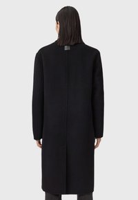 Palton lung negru cu siluetă dreaptă, fabricat dintr-un amestec de lână. Prezintă o cusătură pe spate și un etichetă rectangulară la gât. Design minimalist.