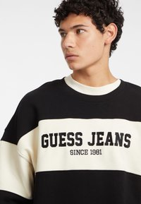 Schwarzer und cremefarbener Pullover mit dem Text "GUESS JEANS". Runder Halsausschnitt, lockere Passform und kontrastierende horizontale Streifen um die Brust.