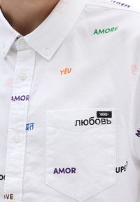 Bílá košile s knoflíky, na které jsou různé fráze v několika jazycích, včetně "любовь" a "AMOR", malé kapsičky a značkového štítku.