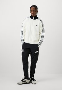 Weißer, flauschiger Zip-Up-Jacke mit schwarzen Streifen an den Ärmeln, kombiniert mit schwarzen Hosen mit deutschem Logo und Adidas-Details. Niedrige Sneakers.