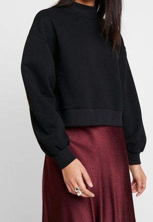 Femme portant un pull court à manches longues noir associé à une jupe en satin bordeaux, montrant une partie du torse et des mains.