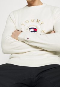 Weißer Sweatshirt mit strukturiertem Stoff, ausgestattet mit einem runden Logo mit "TOMMY" und einem dreifarbigen Emblem. Beinhaltet ein kleines Logo-Detail am Bund.