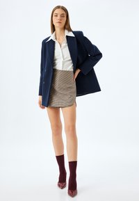 Navyblauwe dubbelgebreide blazer over een witte shirt, gecombineerd met een bruine geruite minirok. Burgondische sokken en glanzende rode puntige hakken.