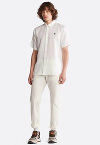 Giovane con i capelli ricci che indossa una camicia bianca a maniche corte, pantaloni chiari, calze bianche e sneakers beige con accenti arancioni.