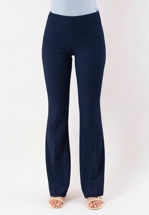 Donna che indossa pantaloni a zampa aderenti blu navy e sandali beige aperti in punta, in piedi contro uno sfondo bianco semplice.