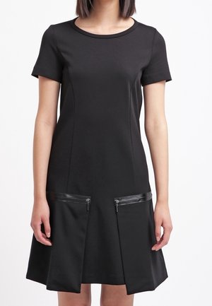 Czarna sukienka z krótkim rękawem, okrągłym dekoltem oraz dwoma przednimi kieszeniami na zamek, noszona przez stojącą osobę na białym tle.
