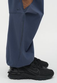 Ναυτικό μπλε αθλητικές παντελόνες με καφέ διακοσμητική λεπτομέρεια, με κομψό σχέδιο και ορατές ραφές, συνδυασμένες με μαύρα αθλητικά παπούτσια Nike.