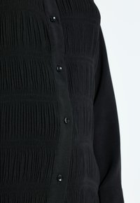 Chemise noire à manches longues avec une texture verticale froncée et des boutons noirs le long de la patte de boutonnage avant.
