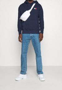 Sudadera marinera con logo, jeans de un azul claro y zapatillas blancas. La sudadera tiene un bolsillo en la parte delantera y está hecha de una tela suave. Bolsa bandolera blanca.