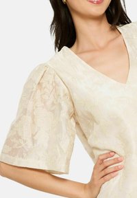 Kreemi värvi kleit lühikeste puhvbliste varrukatega. Omab V-kaelust ja lillemustrit, mis katab kangast, tekstuurse säraefektiga.