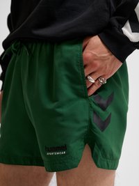 Prawa ręka z srebrnymi pierścieniami w kieszeni zielonych sportowych szortów Hummel noszonych z czarnym topem z długim rękawem.