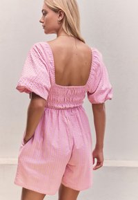 Vaaleanpunainen kuviollinen jumpsuit, jossa on smokkauksella koristeltu selkä, lyhyet hihat ja sivutaskut. Kangas on vaakaraitainen ja siinä on teksturoitu pinta.