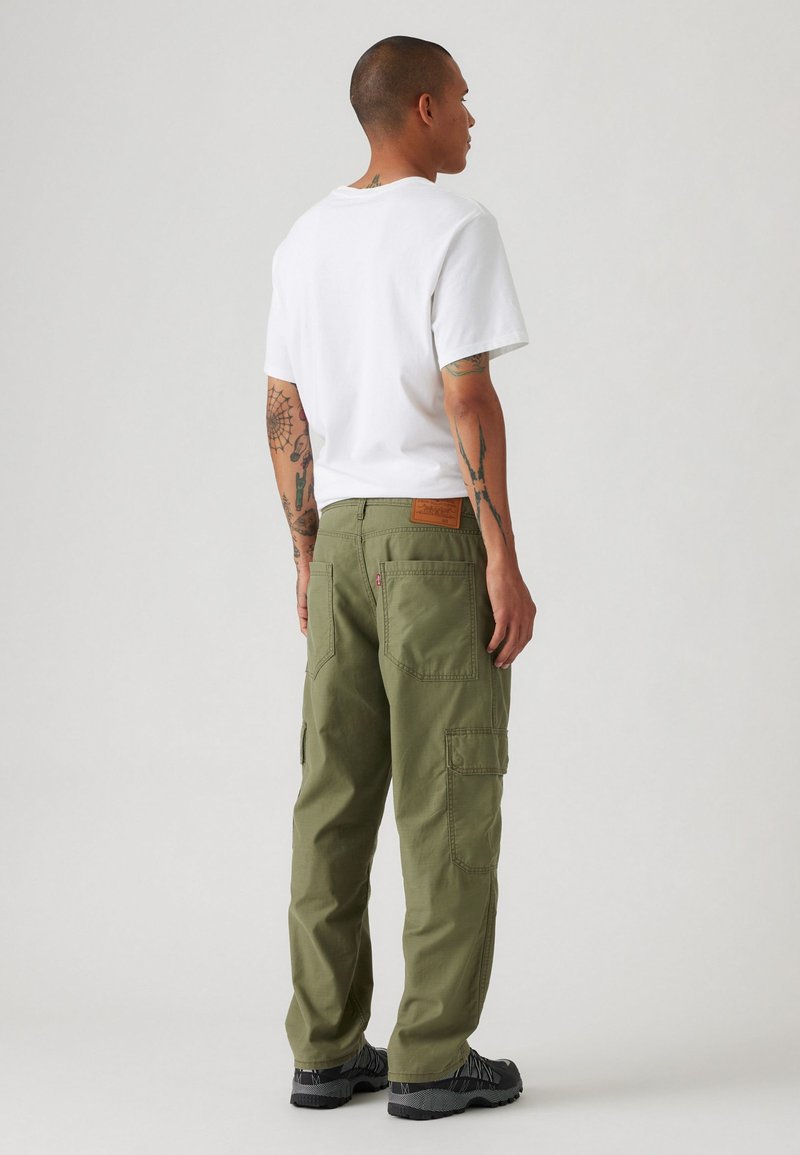 Pantaloni cargo verde oliva con più tasche, in tessuto di cotone, e una patch in pelle del marchio sul retro. Abbinati a una t-shirt bianca semplice.