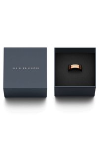 Ruusukultainen sormus esillä tummansinisessä laatikossa, jossa on kultaiset kirjaimet. Pehmeä musta sisustus korostaa kiillotetun sormuksen pintaa.