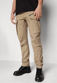 Beige Cargo-Hosen mit mehreren Taschen, Reißverschlussdetails und lockerer Passform, kombiniert mit schwarzen Schnürstiefeln. Texturiertes Material, lässiges Design.