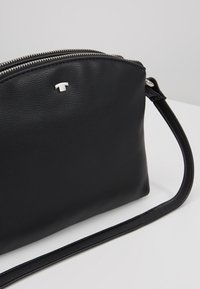 Czarna skórzana torebka na nadgarstek o teksturowanej powierzchni, z srebrnym zamkiem błyskawicznym i małym srebrnym logo z przodu, leżąca na białej powierzchni.