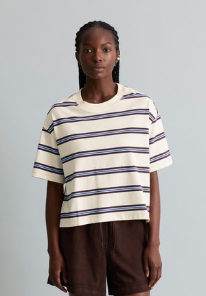Femme aux cheveux tressés portant une chemise crème à manches courtes avec des rayures horizontales bleues et marron et un pantalon brun foncé sur un fond gris.