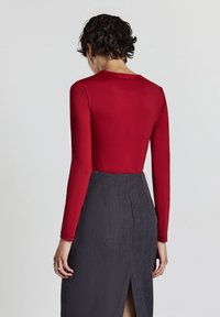 Persona con capelli corti e ricci che indossa un top rosso aderente a maniche lunghe e una gonna grigia a vita alta con spacco sul retro, rivolta all'indietro.