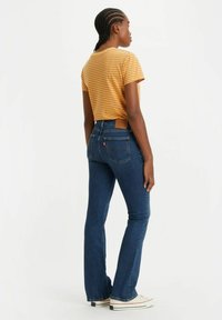 T-shirt a righe color senape con maniche corte, abbinata a jeans aderenti di colore blu scuro. I jeans presentano una tasca posteriore e una patch con il marchio sulla vita.