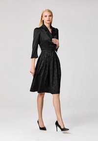 Femme blonde portant une robe noire longueur genoux avec des épaules bouffantes et des manches trois-quarts, associée à des escarpins noirs, se tenant sur fond blanc.