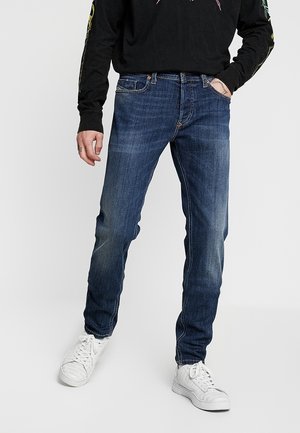 Personne portant un jean slim bleu foncé, des baskets blanches et une chemise noire à manches longues avec des motifs graphiques sur les manches.