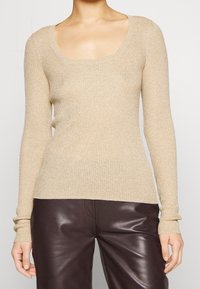 Haut beige côtelé à manches longues avec un col en arrondi, présentant un tissu texturé qui reflète la lumière, associé à un pantalon marron foncé.