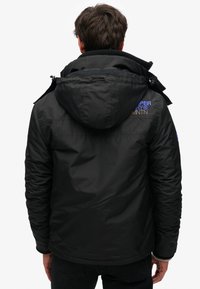 Zwarte geïsoleerde jas met een capuchon, voorzien van een gestructureerde stof, ritssluiting en contrasterende blauwe en tan logo-details op de achterkant.